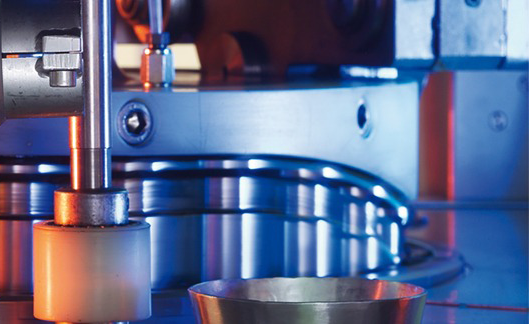 Nahaufnahme einer Industriemaschine mit Metall- und Kunststoffteilen, hervorgehoben durch blaue und orangefarbene Beleuchtung, die einen Prozess der pharmazeutischen Lohnherstellung mit einem kleinen Metallbecher im Vordergrund zeigt. Nahaufnahme einer Industriemaschine mit Metall- und Kunststoffteilen, hervorgehoben durch blaue und orangefarbene Beleuchtung, die einen Prozess der pharmazeutischen Lohnherstellung mit einem kleinen Metallbecher im Vordergrund zeigt.