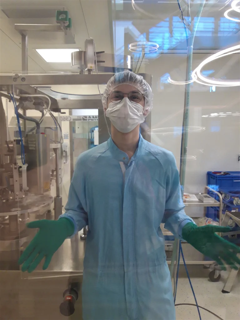 Eine Person in einem Reinraumanzug, mit Maske, Haarnetz und grünen Handschuhen steht in einem Labor oder einer Produktionsstätte hinter einer transparenten Barriere, wobei Maschinen und Geräte im Hintergrund sichtbar sind.