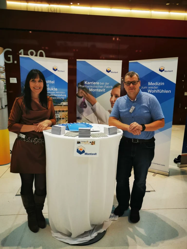 Zwei Personen stehen lächelnd hinter einem runden Tisch mit Broschüren an einem Montavit-Stand. Drei Banner hinter ihnen zeigen Informationen über Arzneimittel, Berufe und Medizin. Die Umgebung scheint eine Indoor-Veranstaltung oder Messe zu sein.