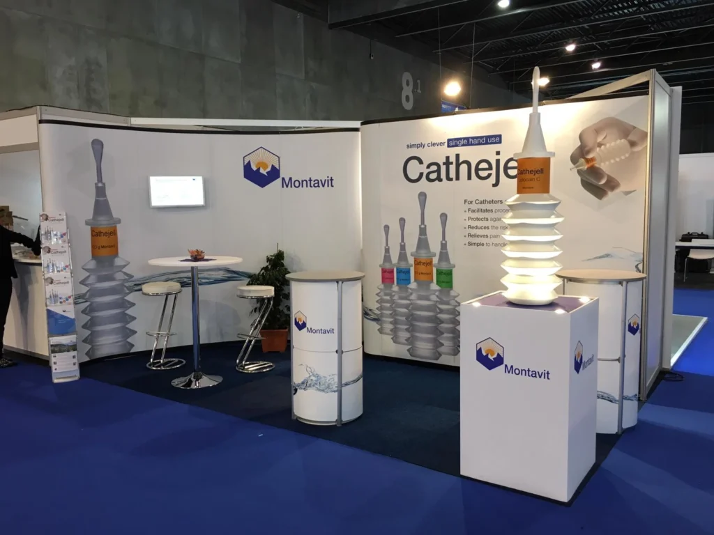 Messestand mit Montavit-Branding, bestehend aus einem großen Catheje-Produktdisplay, Informationsplakaten, einem hohen Tisch mit Stühlen, Produktmustern und einem beleuchteten Werbeständer, alles auf einem blauen Teppich.
