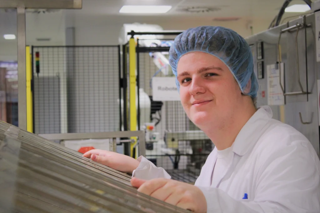 Eine Person in einem weißen Laborkittel und einem blauen Haarnetz steht in einer Industrie- oder Laborumgebung, lächelt und arbeitet mit Geräten.
