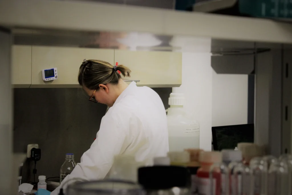 Eine Person in einem weißen Laborkittel arbeitet an einem Labortisch, umgeben von Flaschen und wissenschaftlichen Geräten. Die Person hat ihr Haar zurückgebunden und ist auf eine Aufgabe konzentriert, die sie von hinten durch ein Regal betrachtet.