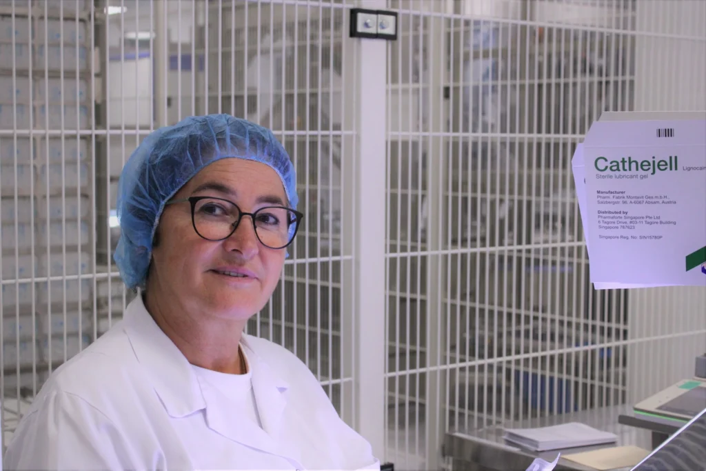 Eine Frau mit Brille, blauem Haarnetz und weißem Laborkittel steht in einem Labor neben einem Schild mit der Aufschrift Cathejell. Sie lächelt und blickt in die Kamera, während im Hintergrund Regale und Geräte zu sehen sind.