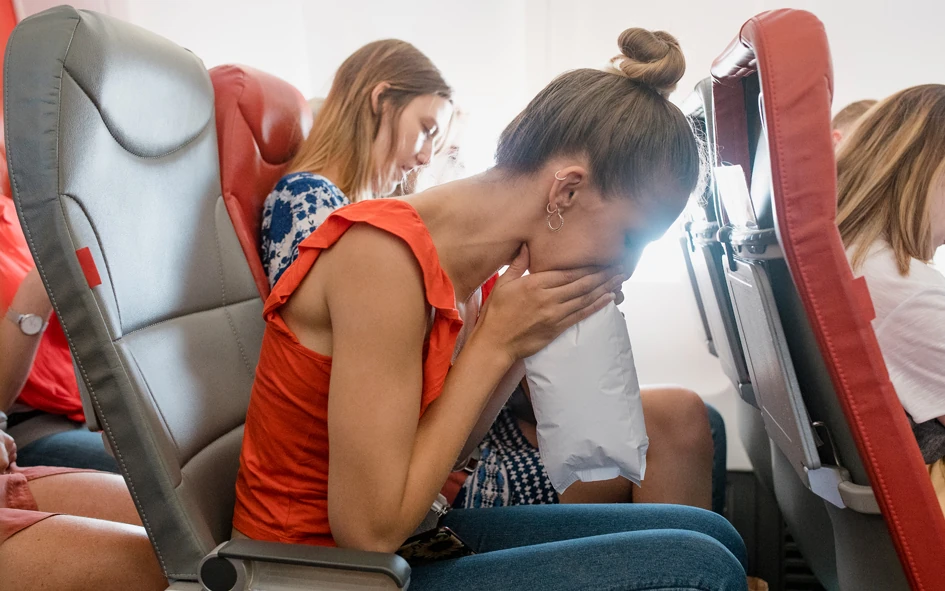 Eine Frau, die auf einem Flugzeugsitz sitzt, lehnt sich nach vorne und atmet in einen Air Sickness Bag, während andere Passagiere in der Nähe sitzen.