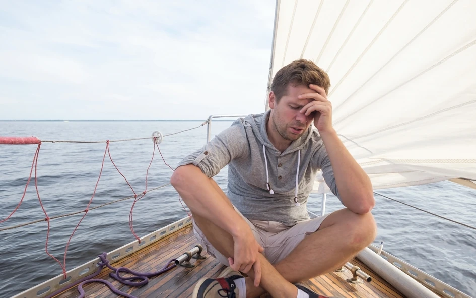 Ein Mann sitzt auf einem Segelboot mit der Hand auf der Stirn und sieht gestresst oder unwohl aus, während er von ruhigem Wasser, dem Segel und den Seilen des Bootes umgeben ist.